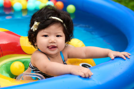 Asian baby girl in swimsuit and float swimming in pool with ballsの写真素材