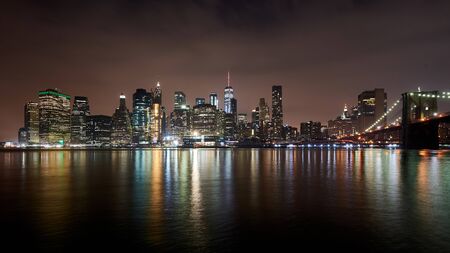 Great night view of the famous skyline of Manhattan downtown district with many skyscrapers and Brooklyn Bridge, whose lights reflects in the water.の写真素材