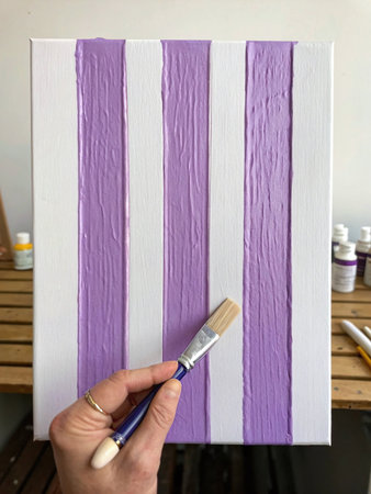 A hand holding a paint brush and painting a vertical purple line on a white canvas frame. AI Generated.の素材