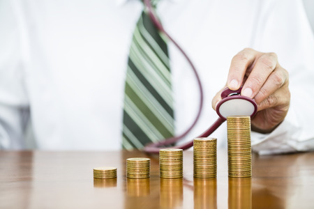 Businessman hand holding stethoscope checking stack of money coins arranged as a graph on wooden table, concept of financial health check and saving moneyの写真素材