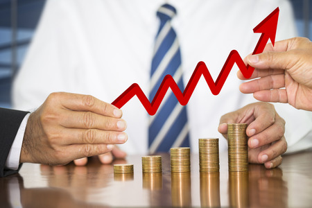 Businessman hand holding a red arrow up over money coin stack arranged as a graph on wooden table and putting money coin, concept of money growth and help to raise fundsの写真素材