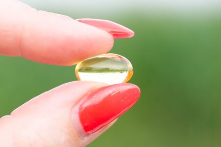 Yellow and transparent pill held between thumb and finger isolated on natural green background. Red nails.の写真素材