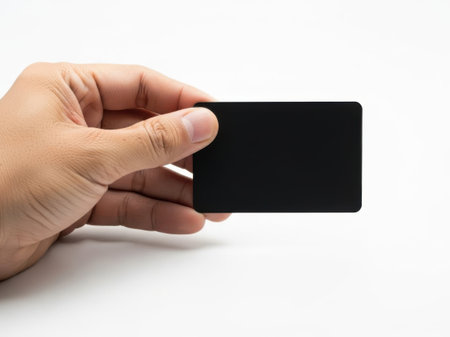 The image shows a hand holding a blank credit card against a plain white background. The card is held horizontally, with the thumb and index finger gripping it from the sides. The hand appears to be in a relaxed position, with the other fingers slightly curled. The background is plain and uncluttered, focusing all attention on the hand and the card. The card itself is black and appears to be made of plastic, with no visible text or design on its surface.の素材