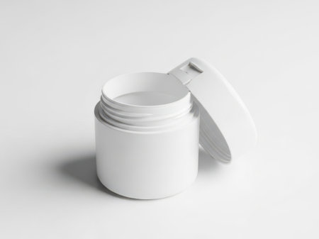 The image depicts a single white cosmetic jar with its lid partially removed, revealing the inside. The jar is cylindrical with a smooth finish and sits on a plain background. The lid is slightly tilted, exposing the open top of the jar. The overall setting is minimalistic, emphasizing the simplicity and cleanliness of the container.の素材