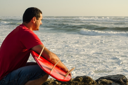 A surfer watching the waves sitting down with his arms around his surfboard.の写真素材