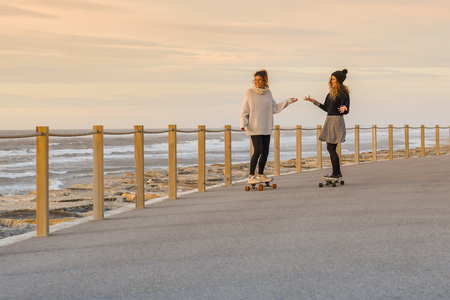 Two female friends playing with skateboard near the beach at sunset.の写真素材