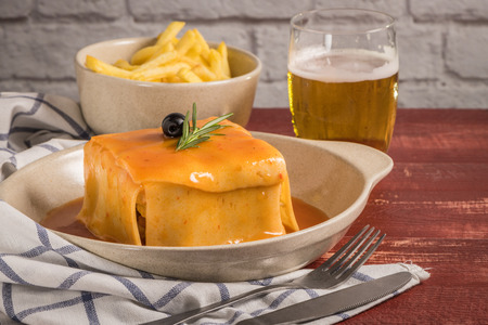 Traditional Portuguese snack food. Francesinha sandwich of bread, cheese, pork, ham, sausages, with tomato beer sauce and French fries. With beer and potatoes. On table.の写真素材