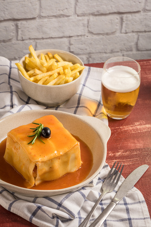 Traditional Portuguese snack food. Francesinha sandwich of bread, cheese, pork, ham, sausages, with tomato beer sauce and French fries. With beer and potatoes. On table.の写真素材