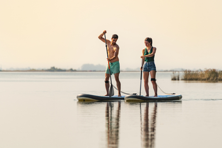 Man and woman stand up paddleboarding on lake. Young couple are doing watersport on lake. Male and female tourists during summer vacation.の写真素材