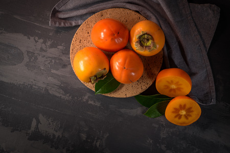 Ripe persimmon fruits in a cork plate on dark kitchen counter top.の写真素材