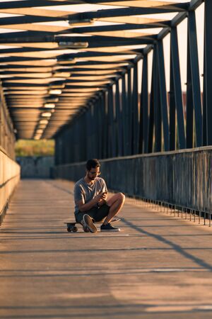 Young man sitting on his skateboard outside on a pedestrian bridge by the river, texting.の写真素材