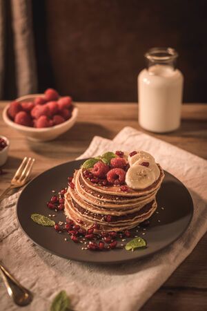 Pancakes with raspberries, banana slices, pomegranate seeds and honey on wooden vintage table.の写真素材