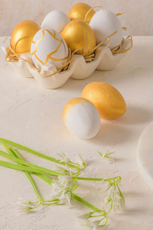 Closeup of golden and white Easter eggs with white flower on bright white wooden table background.