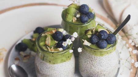 Chia pudding with kiwi, blueberries and coconut slices, three portions in glass jars on a white...