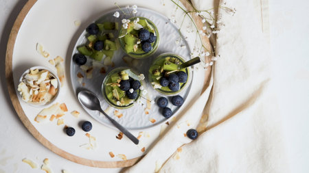 Chia pudding with kiwi, blueberries and coconut slices, three portions in glass jars on a white table.の写真素材