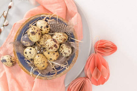 Quail eggs in a ceramic bowl with a peach fuzz pink colored scarf and bird feathers. Wooden table...