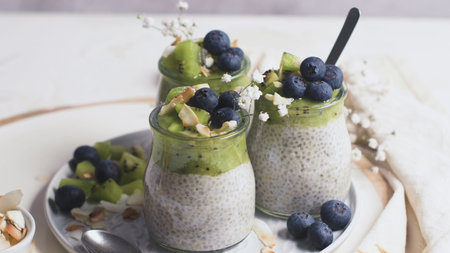 Chia pudding with kiwi, blueberries and coconut slices, three portions in glass jars on a white table.の写真素材