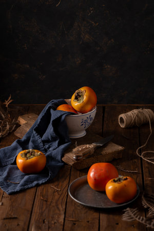 Golden persimmons rest on navy linen against weathered wood. A ceramic bowl and rustic elements create a moody, autumnal scene rich in texture and warmth.の写真素材