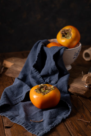 Golden persimmons rest on navy linen against weathered wood. A ceramic bowl and rustic elements create a moody, autumnal scene rich in texture and warmth.の写真素材