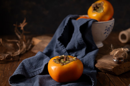 Ripe persimmons on a navy cloth with rustic wooden elements create a cozy autumn scene. Soft lighting highlights the fruit's vibrant color and texture.の写真素材