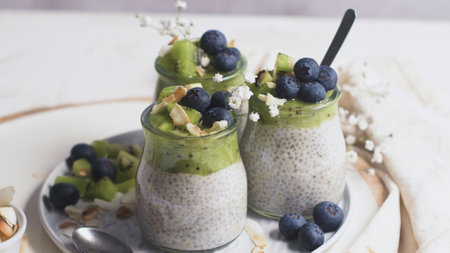 Three jars filled with creamy chia seed pudding, topped with fresh kiwi slices, blueberries, almond flakes, and delicate white flowers.の写真素材