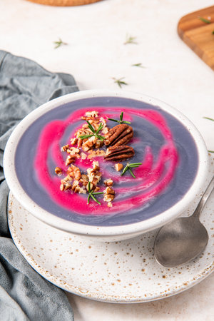 Vibrant purple soup with a pink swirl, garnished with chopped pecans and fresh rosemary, served in a speckled white bowl on a matching plate with a spoonの写真素材