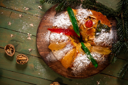A traditional holiday sweet bread dusted with powdered sugar and adorned with candied fruits and green citrus peel, served on a rustic wooden board with pine branchesの写真素材