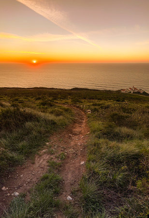 A breathtaking sunset over the Atlantic Ocean near Farol do Cabo Mondego in Figueira da Foz, Portugal, with a dirt path leading through a grassy landscape.の写真素材