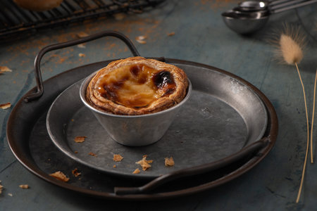 A single Portuguese custard tart in a metal baking tin rests on a round metal tray with measuring...