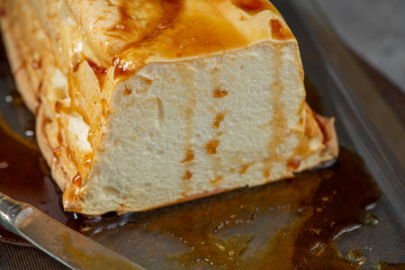 A close-up showcases a substantial slice of molotof, a classic Portuguese caramelized egg white pudding, generously drizzled with a rich amber syrup on a dark surfaceの写真素材