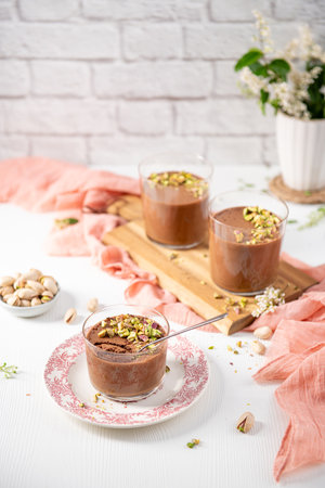 Three servings of rich chocolate dessert, topped with vibrant pistachios, are artfully arranged on a white table with a pink cloth and a bowl of nutsの写真素材