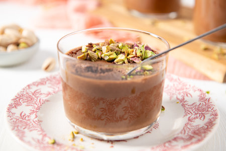 A delightful chocolate dessert, topped with vibrant green pistachios, sits in a clear glass on a decorative plate, ready to be enjoyed with a spoonの写真素材