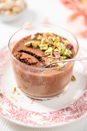 A single serving of delectable chocolate dessert, topped with chopped pistachios, is presented in a clear glass with a spoon on a decorative white and red plateの写真素材
