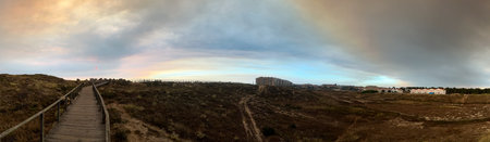 A wide coastal view with a boardwalk leading through sandy terrain towards distant buildings under a...
