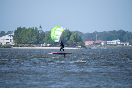 A middle-aged man in a wetsuit glides on a hydrofoil board with a green and white kite, with houses...