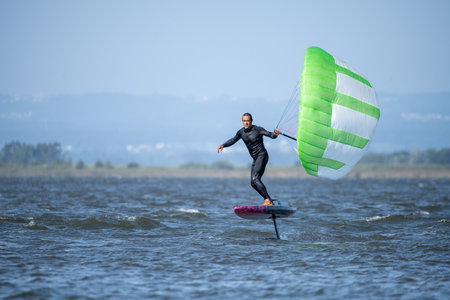A middle-aged man in a wetsuit glides on a hydrofoil board with a green and white kite, looking at...