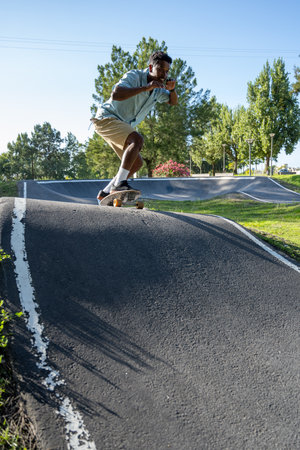 A young Black man actively rides his skateboard down a steep, asphalt ramp at a pump track on a bright, sunny day. He is crouched low and focused, using his body to navigate the incline with power and control. The image captures the dynamic energy of the sport set against a backdrop of lush green grass and trees.の写真素材