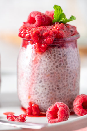 A detailed, eye-level close-up of a single glass jar of chia seed pudding, topped with a generous layer of homemade raspberry compote, fresh raspberries, and a mint leaf. The healthy, vibrant breakfast is presented on a white plate, with a focus on the pudding's unique texture and the rich, red colors of the fruit, set against a bright, modern backdrop.の写真素材