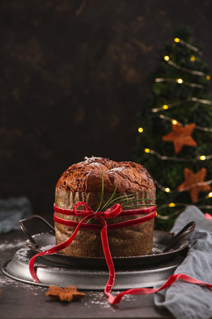 A festive Panettone, the classic Italian holiday cake, is presented on a rustic metal tray, tied with a rich red velvet ribbon and lightly dusted with powdered sugar. The vertical image is styled with a dark, moody background and a small, decorated Christmas tree with warm yellow lights and star ornaments.の写真素材