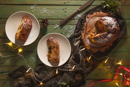 An overhead flat lay shows a whole, sliced, and sugar-dusted panettone on a rustic wood board next to two served slices on white plates. The dark green wooden background is heavily styled with fairy lights, pinecones, scattered sugar, and a knife, creating a moody, rich, and festive Christmas ambiance.の写真素材