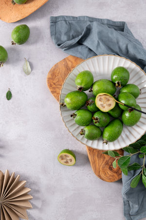 A vibrant vertical shot of fresh, green feijoa fruits arranged on a white fluted plate. One of the fruits is cut in half, revealing its pale, golden interior, with a small black spoon resting inside. The composition is set on a neutral gray counter with a rustic wooden board, a blue linen napkin, and a decorative paper fan visible, creating an attractive and natural aesthetic.の写真素材