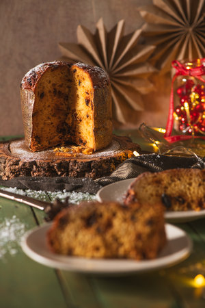 A vertical shot of a sliced, sugar-dusted panettone on a rustic wood slice with two slices served on plates in the soft-focus foreground. The warm, dark, and moody lighting, along with Christmas decorations, enhances the festive, cozy, and traditional holiday dessert scene.の写真素材