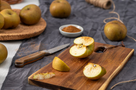 A rustic still life scene captures sliced green and yellow apples on a wooden cutting board, lightly dusted with cinnamon. A kitchen knife, a bowl of cinnamon, and whole apples are scattered on a dark gray cloth, suggesting a cozy autumn baking or cooking preparation.の写真素材