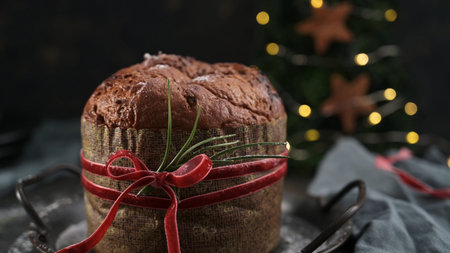 A warm, close-up studio shot of a traditional Italian Panettone cake, presented as a gift wrapped in patterned paper and tied with a red velvet ribbon and rosemary sprig. The dark, moody background features blurred Christmas tree lights and wooden star decorations, creating an inviting and rustic holiday atmosphere.の写真素材