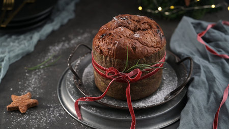 A traditional Italian Panettone cake is centered on stacked metal serving platters, wrapped in a patterned sleeve and tied with a red velvet ribbon and rosemary sprig. The dark, moody setting is scattered with powdered sugar, creating a rustic holiday atmosphere, with a blurry string of Christmas lights visible in the background.の写真素材