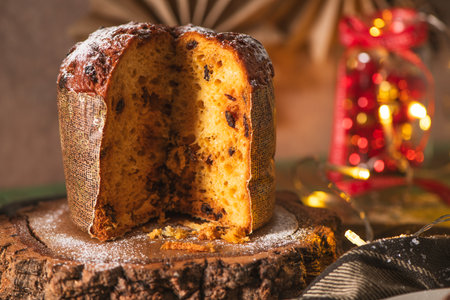 A round, traditional panettone with chocolate chips is sliced to reveal its moist interior, dusted with powdered sugar, and displayed on a rustic wooden slice. The warm, inviting chiaroscuro lighting emphasizes the cozy, winter holiday mood, complemented by blurred background string lights.の写真素材