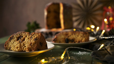 Close-up of moist panettone slices on white plates, illuminated by the warm glow of string lights against a dark, rustic wooden table. The festive, cozy mood is set by the shallow depth of field and soft background holiday decor.の写真素材