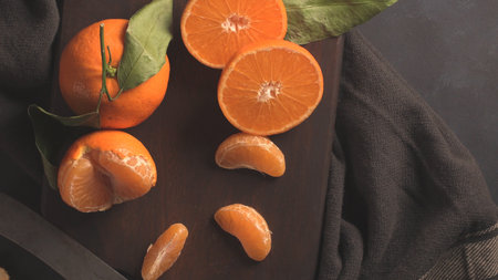 An overhead, dark food photography shot featuring vibrant mandarins arranged on a rustic, dark wooden cutting board surrounded by a gray cloth and a knife blade. The composition includes whole fruit with leaves, halves, and individual peeled segments, emphasizing the fresh, rustic quality of the citrus.の写真素材