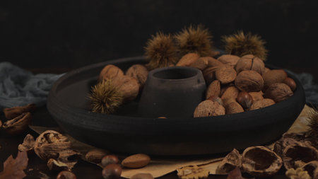 A moody, low-key still life featuring walnuts, almonds, and hazelnuts in a matte black bowl. Warm...