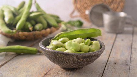 A bowl of bright green fava beans sits on a weathered wooden surface. Soft natural lighting...